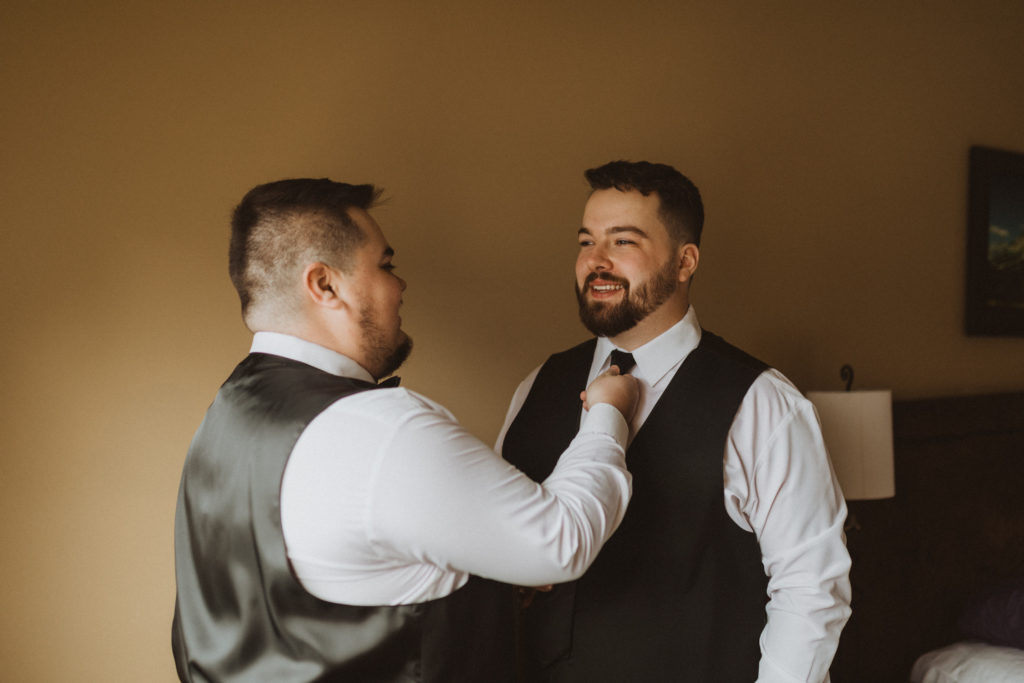Groom gets ready with best man morning of his elopement day. Portrait of groomsman tying groom's tie. 
