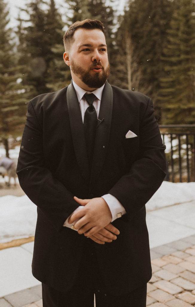 groom's reaction seeing his bride walk down the aisle. Groom is wearing a black tux and it is snowing during their wedding ceremony in Canmore, Alberta.