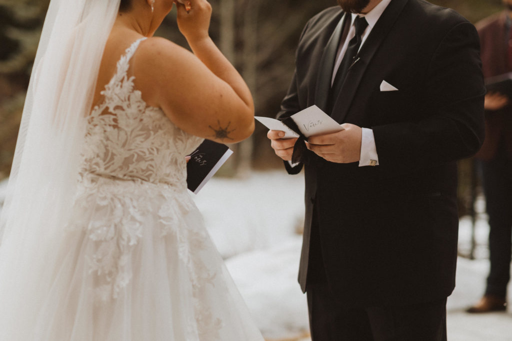 Bride and groom reading personal vows to each other from custom handmade vow books. Bride wearing lace ballgown wedding dress, and groom wearing black suit with square cufflinks.