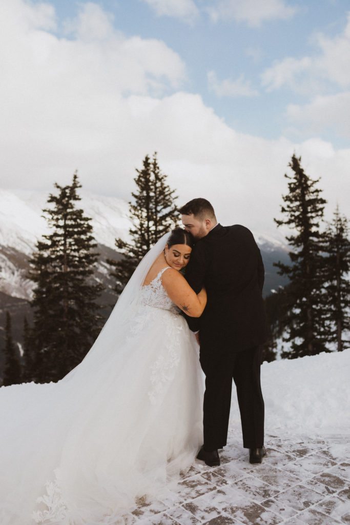 Bride and groom portrait standing side by side all wrapped up on his arm and kissing her on the head. We took their elopement portraits on top of Sulphur mountain up the Banff Gondola 