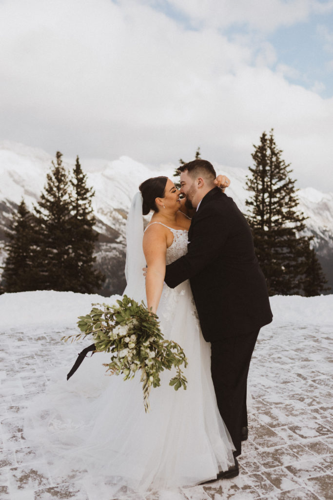 Banff gondola elopement portraits on top of sulphur mountain with green bouquet from Always Sunny Design. Bride and groom about to kiss surrounded by the snow peaked rocky mountains.