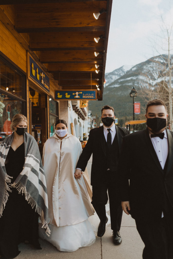 Bride, groom, maid of honour, and best man walking through the town of Banff on elopement day and wearing masks because they got married during the covid-19 pandemic.