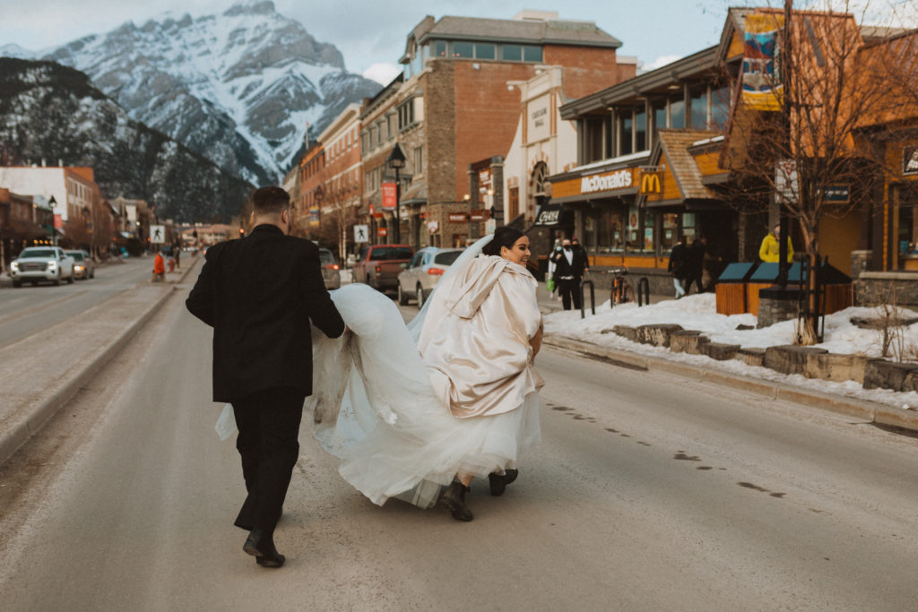 Bride and groom running across the street in the town of Banff to McDonalds to spend their dinner eating fries and a Big Mac.