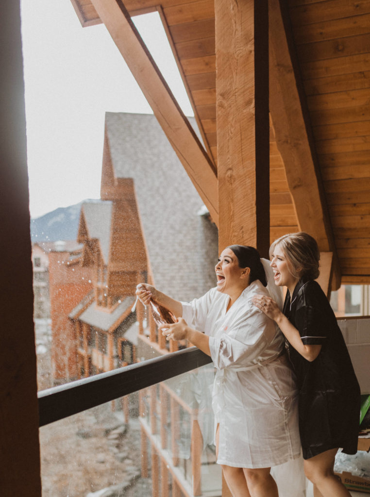 Bride and maid of honour pop champagne off the balcony while getting ready at Solara Resort in Canmore, Alberta.