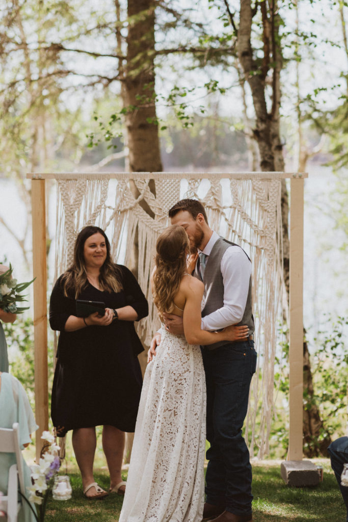 Bride and groom first kiss during ceremony. Macrame arbor as backdrop. Overlooking the water. Saskatchewan elopement photographer. Bride was wearing a lace wedding dress with tan lining from Lulus and groom was wearing blue tie with grey vest.