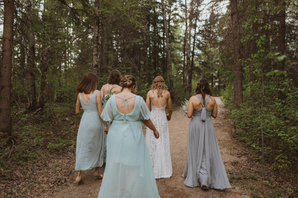 Bridesmaids walking through forest. Flora Bora Yurt Elopement. Wedding party portraits. Bridesmaids wearing mismatched blue bridesmaid dresses with bride in a lace wedding dress from Lulus. Groomsmen wore jeans, cowboy boots, and suspenders from Lamlees. Saskatchewan elopement photographer. Creative bridal party photo ideas.