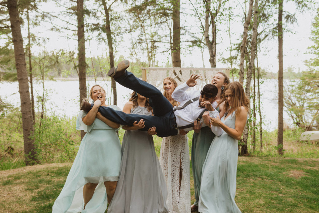 Bridesmaids holding groomsman for photo. Flora Bora Yurt Elopement. Wedding party portraits. Bridesmaids wearing mismatched blue bridesmaid dresses with bride in a lace wedding dress from Lulus. Groomsmen wore jeans, cowboy boots, and suspenders from Lamlees. Saskatchewan elopement photographer. Creative bridal party photo ideas.