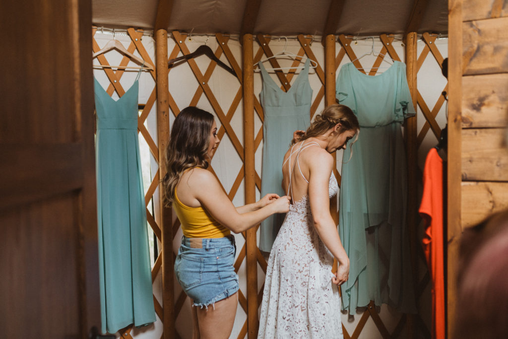 Bride putting on dress with the help of a bridesmaid inside a yurt morning of her intimate wedding. Getting ready photos. Intimate wedding at flora bora.