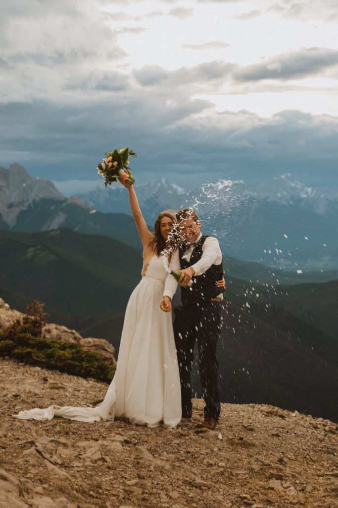 these are all the reasons why you should elope in Jasper National Park. Bride and groom popping a bottle of champagne on top of a mountain in Jasper
