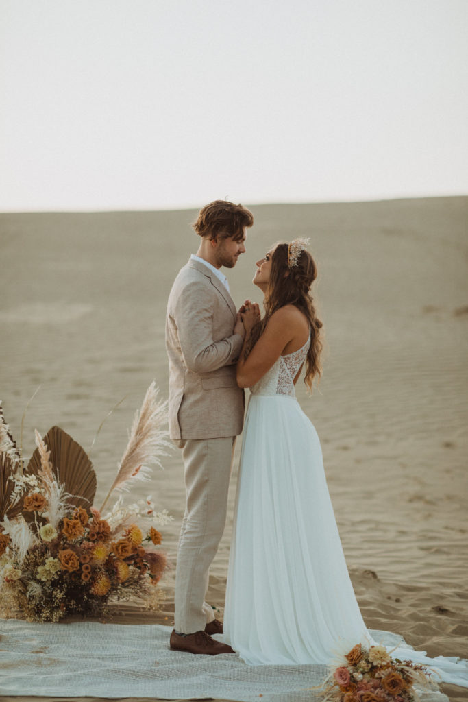 boho elopement ceremony setup, rose toned floral installation, couple standing on rug, bride in flowy dress from Exquisite, groom in tan linen suit