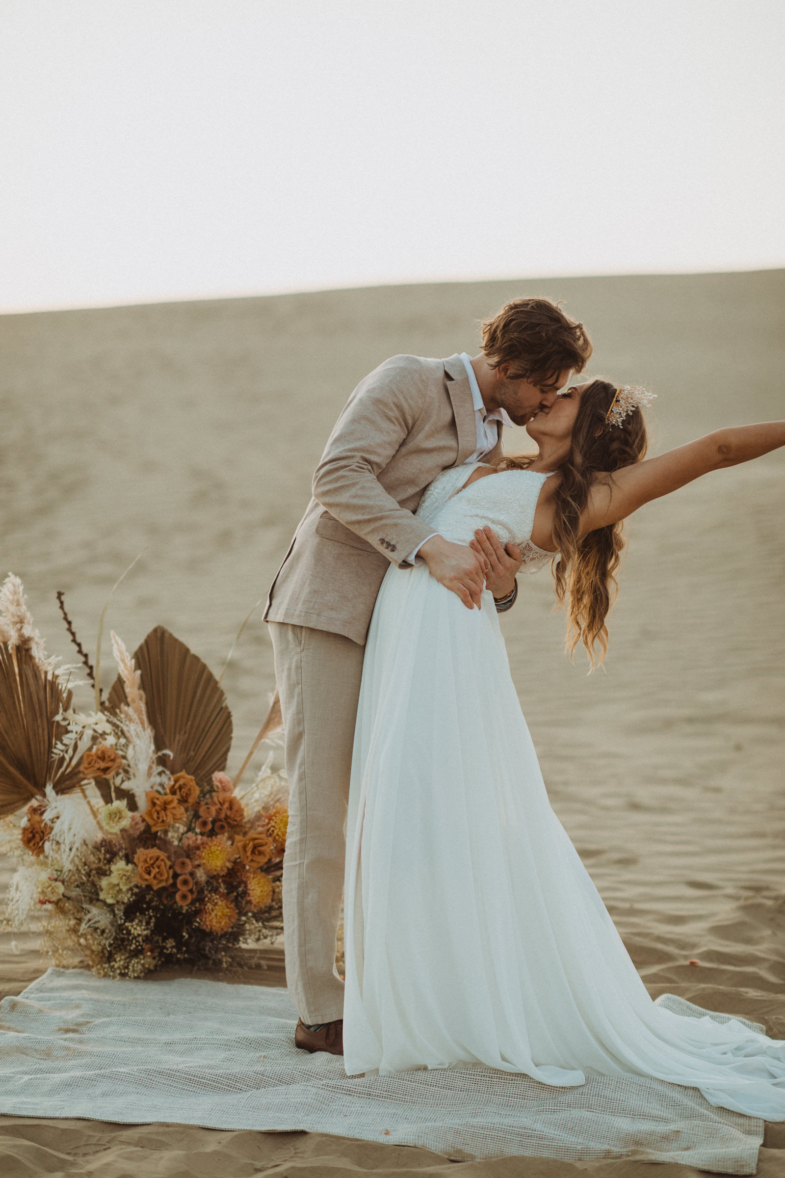 boho elopement ceremony first kiss. rose toned floral installation, couple standing on rug, bride in flowy dress from Exquisite, groom in tan linen suit