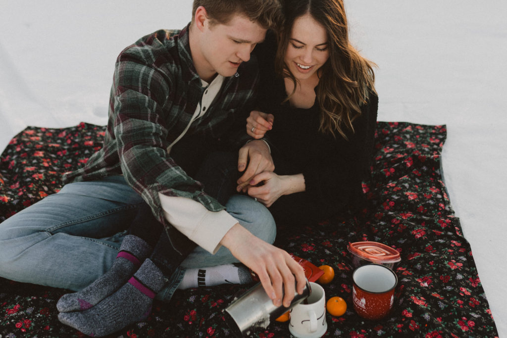 couple enjoying a picnic in the snow with hot chocolate and oranges and a cute blanket because they just got engaged over the holidays and are celebrating
