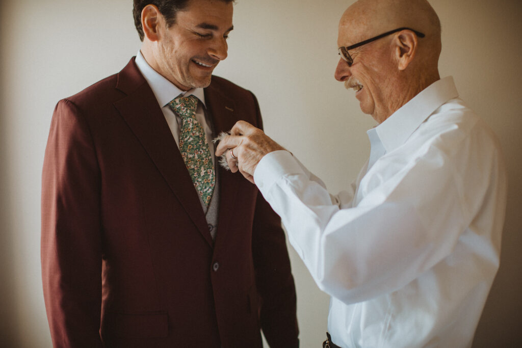 groom getting ready in burgundy suit and green floral tie morning of elopement in an airbnb in Canmore, Alberta