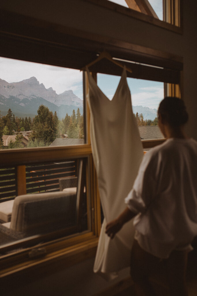 An Airbnb elopement in Alberta captured the excitement of the special day, with the bride and groom getting ready for the ceremony in the cozy, sunlit interior of their rental. The bride stands before a large window overlooking a stunning mountain vista, slipping into her elegant wedding gown. The warm, inviting space serves as the perfect backdrop for the couple's intimate pre-ceremony moments.