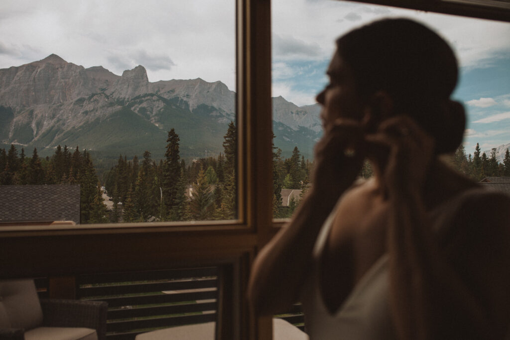bride putting on earrings getting ready for elopement inside a rented airbnb in Canmore, Alberta looking out at the mountains